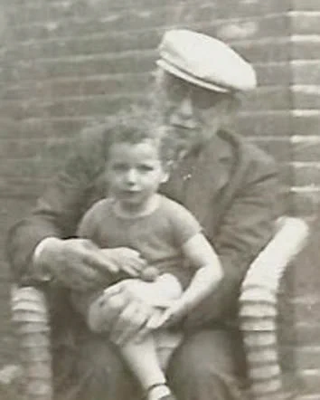 Vintage black and white photo of a child sitting on the lap of an adult wearing a captain's hat, both seated against a brick wall.
