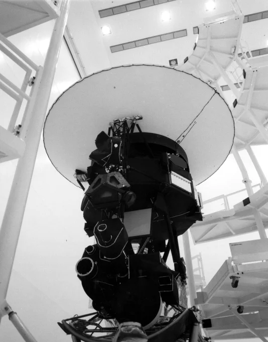 This black and white photograph shows Voyager 1 being encapsulated at Kennedy Space Center ahead of launch. The image is taken from a low angle, looking up at Voyager against a background of clean room scaffolding and infrastructure. 