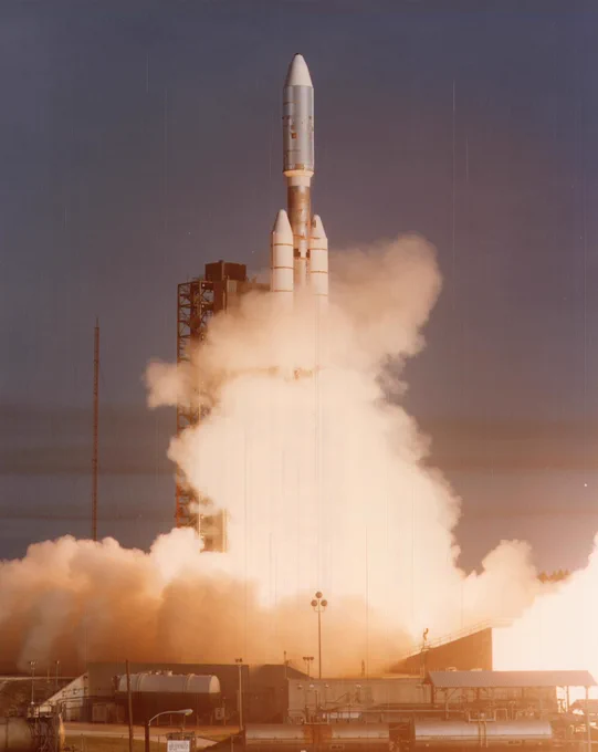 This color photograph shows Voyager 1 launching atop its Titan/Centaur-6 launch vehicle from the Kennedy Space Center on September 5, 1977. Near the top of the frame, the tip of the rocket is visible. Below, the rocket and launchpad is obscured by billowing white clouds of gas and exhaust. The photograph has a grainy vintage filmic quality, with the brightest colors appearing yellowed and the shadows appearing lavender. 