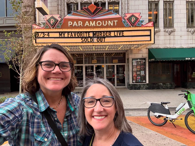Two smiling women taking a selfie in front of Paramount Theater in downtown Denver with a theater sign that reads “My Favorite Murder live — SOLD OUT”