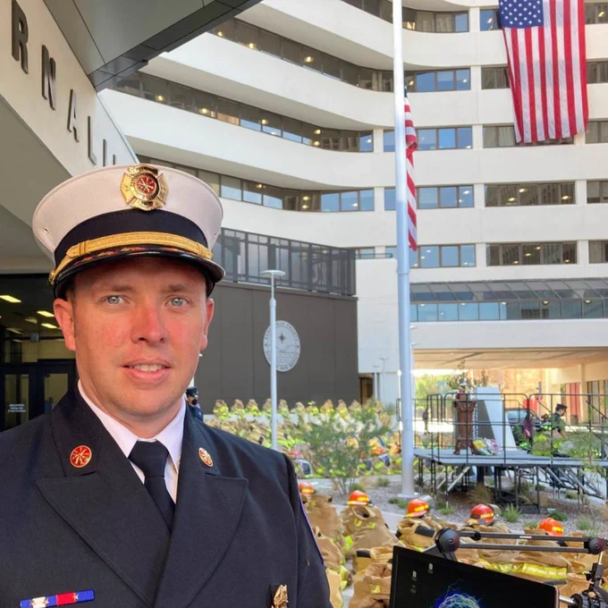 A firefighter in formal uniform stands in front of an American flag and a courtyard with arranged helmets.