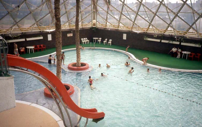 Digitised 35mm colour slide showing a view of a swimming pool from above. Palm trees are planted in the middle and around it. A red slide is in the foreground and people are swimming.