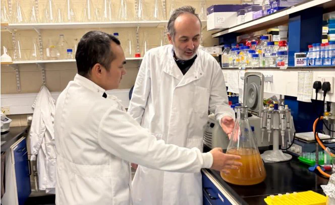 Two scientists in white lab coats examine a large flask containing an orange-brown liquid in a laboratory. The lab is equipped with shelves holding various bottles and containers, and the countertop is cluttered with lab equipment and test tubes.