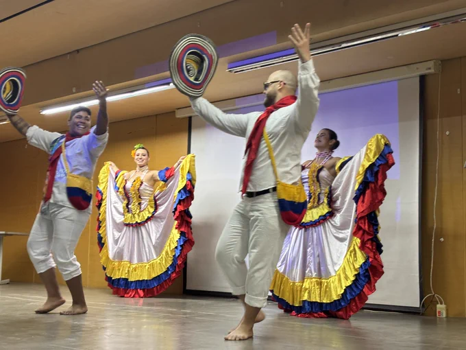 Un grupo de baile. Llevan vestidos coloridos: los hombres con sombrero y vestimenta blanca, las mujeres con falda larga con la bandera colombiana. Están terminando su baile, levantando sus brazos hacia el público.