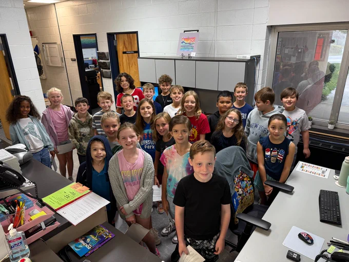 23 fourth graders stand in the front office and smile big for the camera 