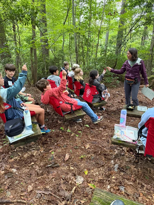 group of fifth grade students sitting in an outdoor learning area in red chairs. they have their hands raised and are facing towards a mountain roots guide who is teaching a lesson 