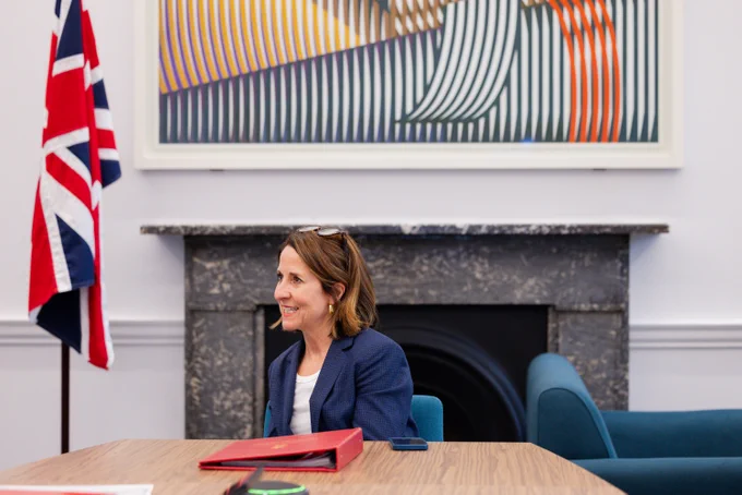 Secretary of State for Science, Innovation and Technology Liz Kendall sitting at a table in the department with a notebook and pen, in a room with a Union Jack flag and a vibrant abstract painting on the wall.