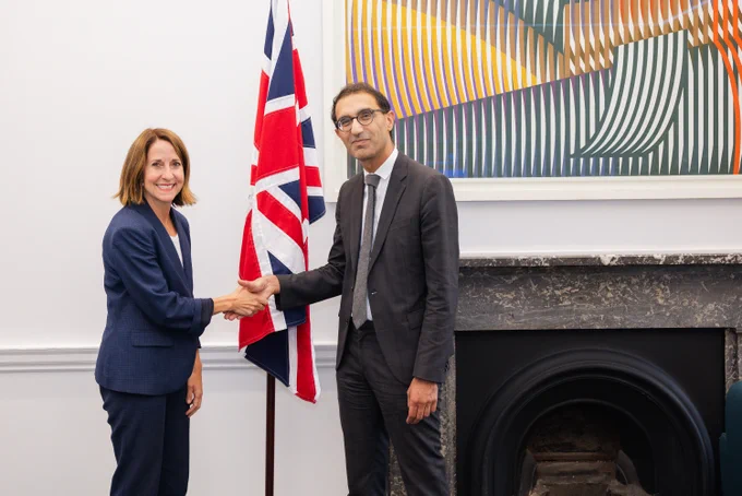 Secretary of State for Science, Technology and Innovation Liz Kendall meets Permanent Secretary Emran Mian in the department, they shake hands in front of a UK flag and an abstract art piece, inside a room with a fireplace.
