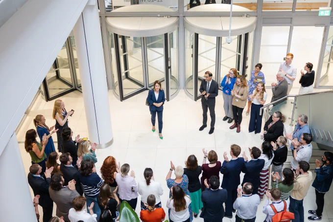 Secretary of State for Science, Technology and Innovation Liz Kendall arrives at the department to meet colleagues, surrounded by a group of people gathered in a modern lobby, listening attentively to her speaking and standing in the center.