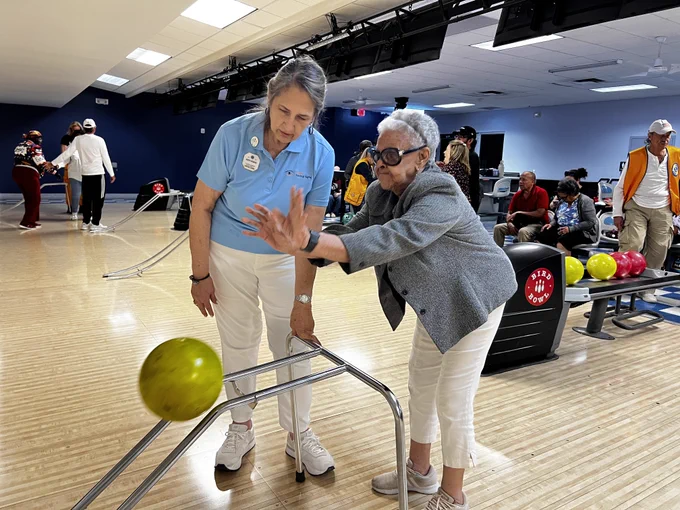 A photo of two people at a bowling alley, in front of a lane, with one person guiding the other through the process of throwing the bowling ball. 