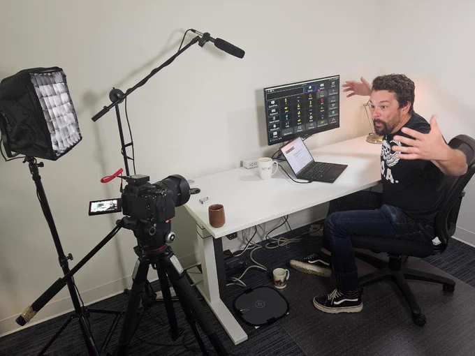 A man sits at a desk in front of a computer screen. He faces a video camera, his arms open wide.