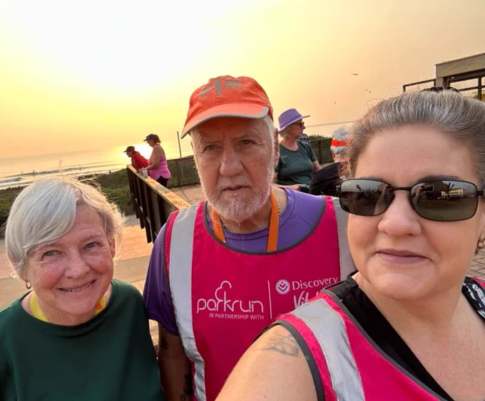 Two volunteers wearing pink vest smiling with a parkrunner in a sunrise selfie.