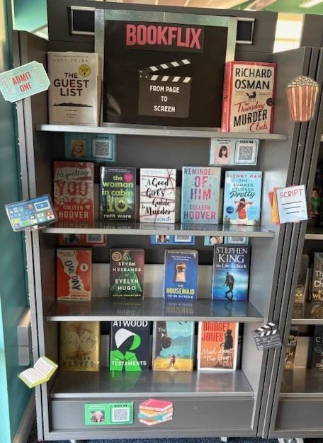 Library display titled “BOOKFLIX: From Page to Screen,” featuring books adapted into movies or TV shows. Three shelves hold titles like The Guest List, It Ends With Us, and The Handmaid’s Tale. Movie-themed decorations include popcorn, clapperboards, and ticket stubs.