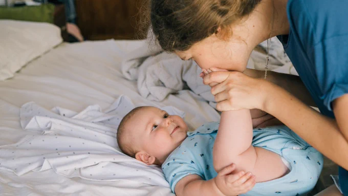 A mum kissing her baby's foot whilst the baby is lying on a bed.