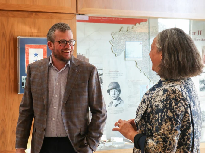 A man wearing a brown blazer and a white button down speaks with a woman wearing a floral jacket in front of a display case.