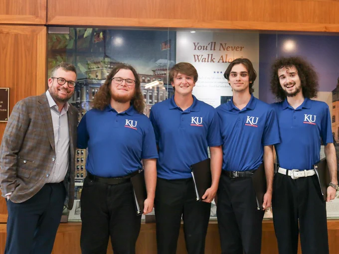 Five individuals posing for a photo in front of the "You'll Never Walk Alone" display at the Dole Institute of Politics, with four of them wearing shirts with "KU" logos.