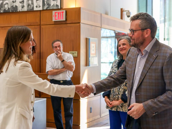 Two individuals shaking hands in a professional setting, with onlookers smiling in the background, beneath a wall of portrait photographs.