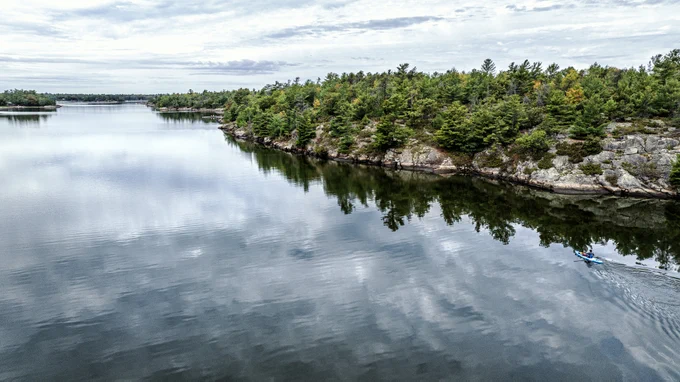 A serene aerial view of a calm lake surrounded by lush green trees and rocky cliffs. The water reflects the sky and the surrounding landscape, creating a mirror-like effect. A lone kayaker paddles along the right side of the image, leaving a small wake behind. The sky is partly cloudy, adding to the tranquil atmosphere of the scene.