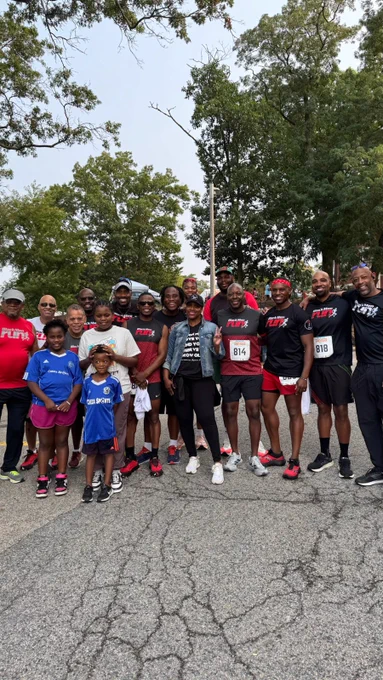 Rep. Pressley, in a hat and jean jacket, holds up a peace sign while surrounded by runners in athletic gear, some of whom are wearing running bibs and shirts that say “Black Men Run.”