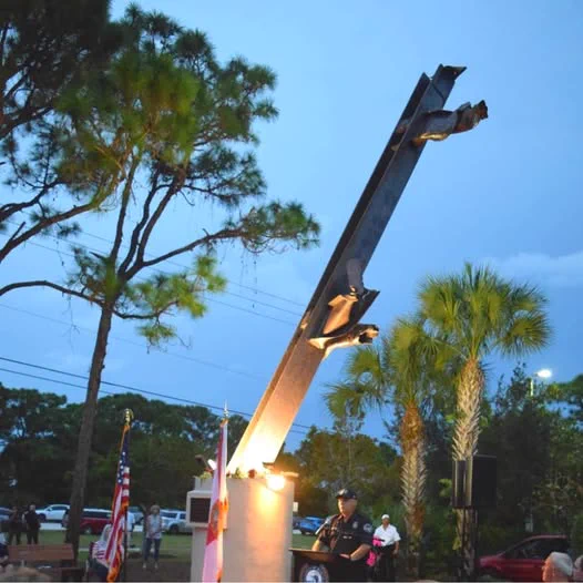 An individual in a military uniform stands at attention beside the flag of the United States at a 9/11 memorial, featuring a large metal beam from the World Trade Center, lit by spotlights at dusk with trees in the background.