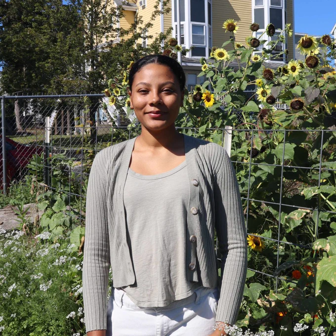 Fatimah stands in front of sun flowers on a sunny day wearing an olive green blouse. 
