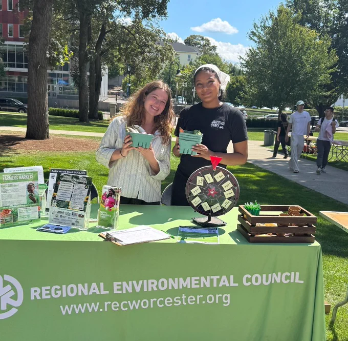 Zoe and Fatimah stand smiling and holding produce in front of a table with a green tablecloth that reads "Regional Environmental Council" on a sunny day at Clark University. The table is covered in flyers, a  prize wheel, and prizes. 