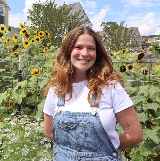 Zoe smiles in front of sunflowers on a sunny day wearing a white t shirt and denim overalls. 
