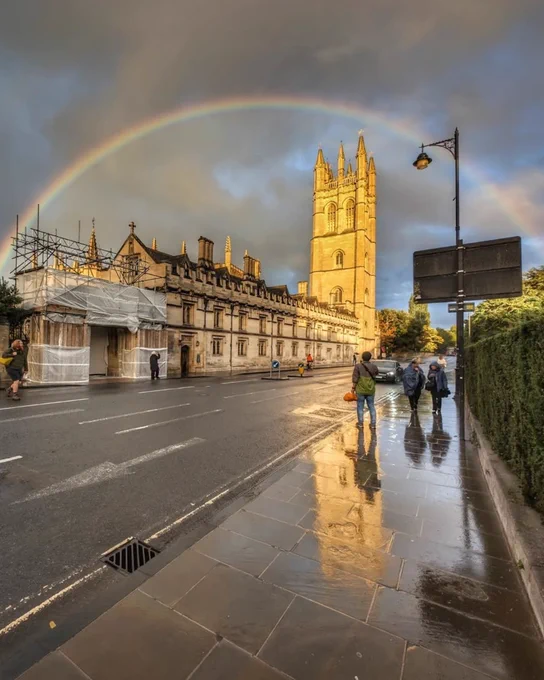 Rainbow arching over Magdalen College in Oxford, with wet pavement reflecting the scene and pedestrians walking by.