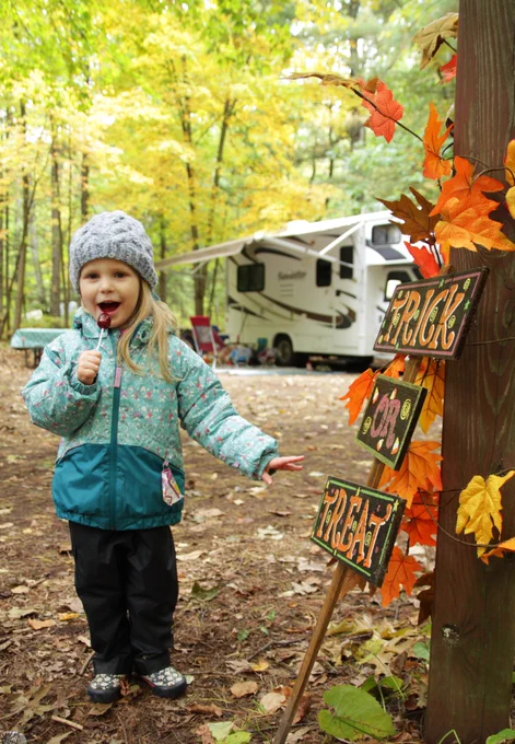 A child wearing a winter hat and jacket stands in a wooded campsite holding a lollipop. An RV is parked among the trees, and a wooden post decorated with autumn leaves displays three signs reading 'Trick,' 'Or,' and 'Treat.' Fallen leaves cover the ground