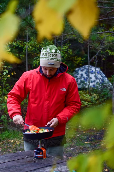 A person in a red jacket and a beanie labeled 'PARKS' cooks vegetables in a pan over a portable camping stove. They are outdoors with a tent and green foliage in the background.