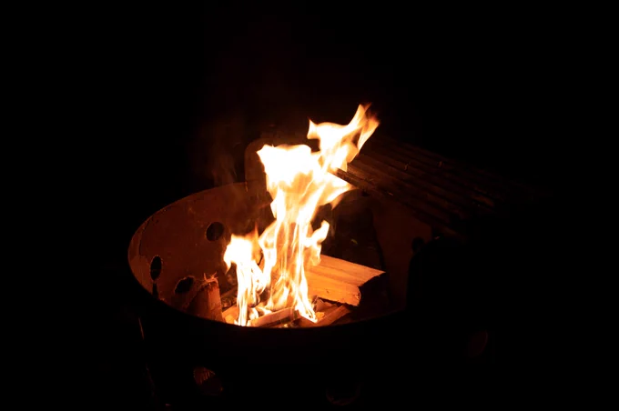 A campfire burning brightly in a metal fire pit with circular holes around its sides, set against a dark background that emphasizes the glow of the flames