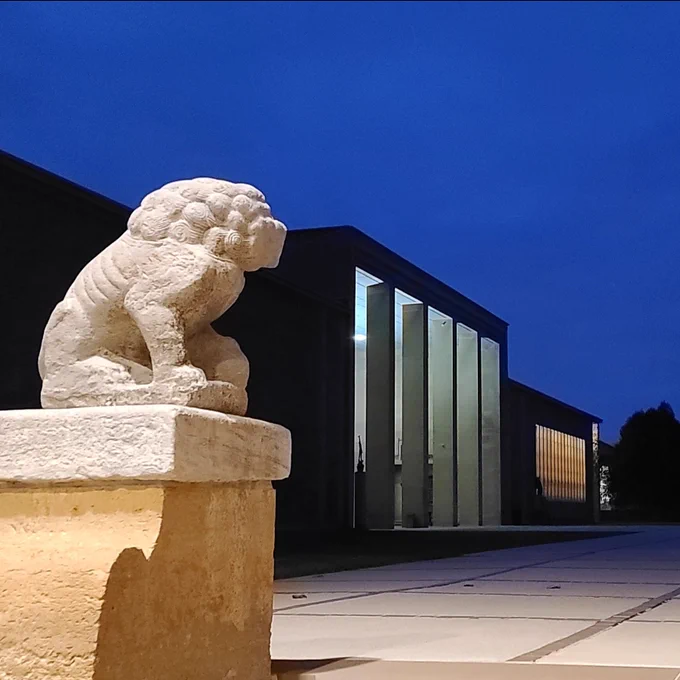 A stone lion statue stands guard at the entrance of a modern building with illuminated vertical windows under a twilight sky.