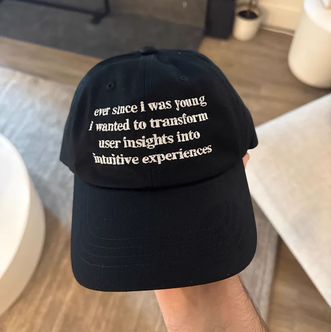Photo of a black hat with white text on the front that says "ever since i was young i wanted to transform user insights into intuitive experiences" 