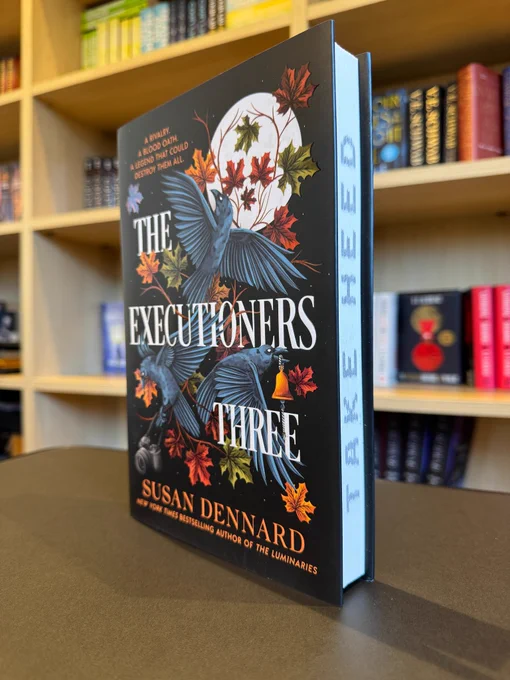 A copy of The Executioners Three by Susan Dennard standing in front of a bunch of bookshelves filled with TPG books.