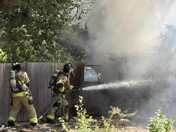 Firefighters from the Bernalillo County Fire Department are actively extinguishing a house fire, with water being sprayed towards the smoke and debris behind a wooden fence.
