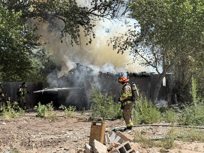A firefighter is extinguishing a fire at a residential building, surrounded by trees and debris.