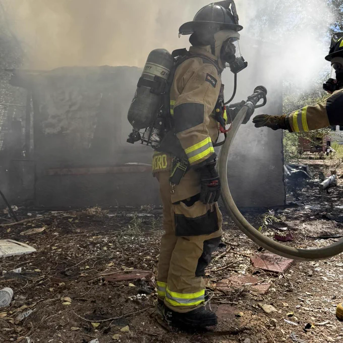 A firefighter wearing full gear, including a helmet, air tank, and reflective stripes, is holding a hose with smoke in the background.