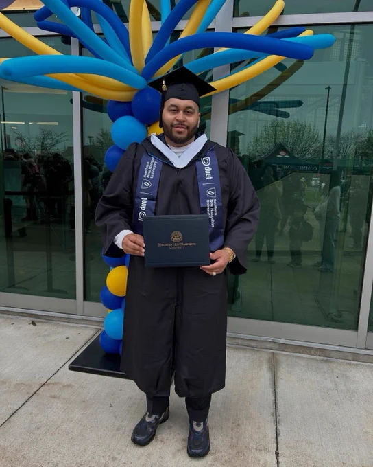 SNHU Graduate stands with diploma in cap and gown in front of balloons. 