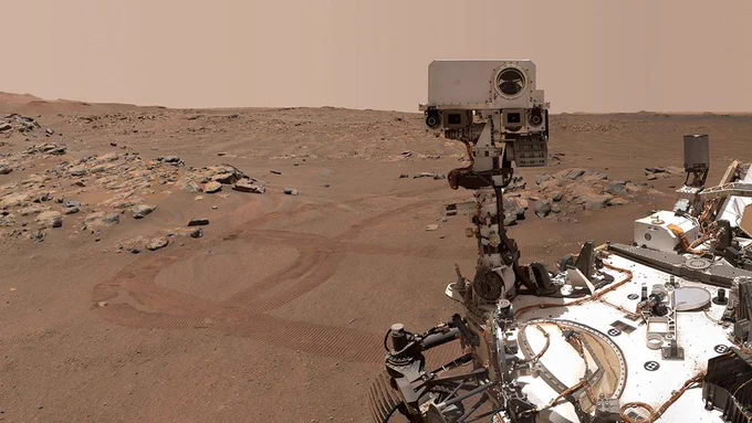 A selfie by the Perseverance rover on Mars. The rover's robotic head is facing the camera. In the background, a rocky, sandy desert landscape, with some fo the rover's own tracks visible on the ground.