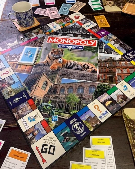 Monopoly game Chester edition. Laid out in play. Tiger to centre of board, Go square to bottom. Diamond shape. Mug and money in background, property cards to foreground.