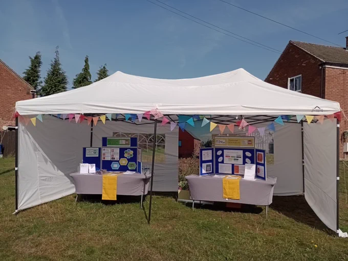 A gazebo decorated with colourful bunting on some grass outside houses. There are display boards with posters and flyers on tables inside.