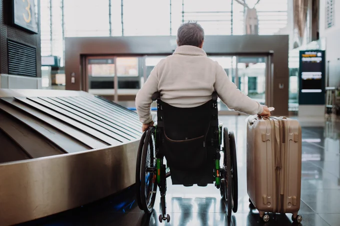 A wide shot photograph of an older white and male wheelchair user with grey hair in an airport foyer. He is sat next to a baggage conveyor belt and has his right arm stretched out, gripping his luggage.