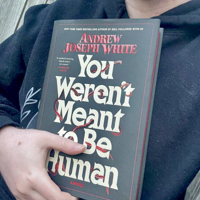 A pale hand holding a copy of YOU WERENT MEANT TO BE HUMAN by Andrew Joseph White against a black sweatshirt.
