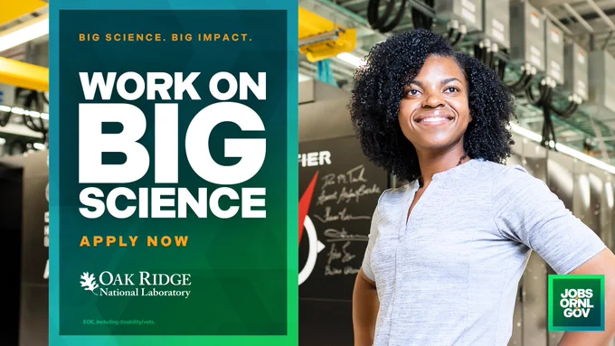 A person smiling confidently in a laboratory, standing next to a poster that reads "Work on BIG SCIENCE. APPLY NOW" with the Oak Ridge National Laboratory logo and the text "Jobs Only Gov.