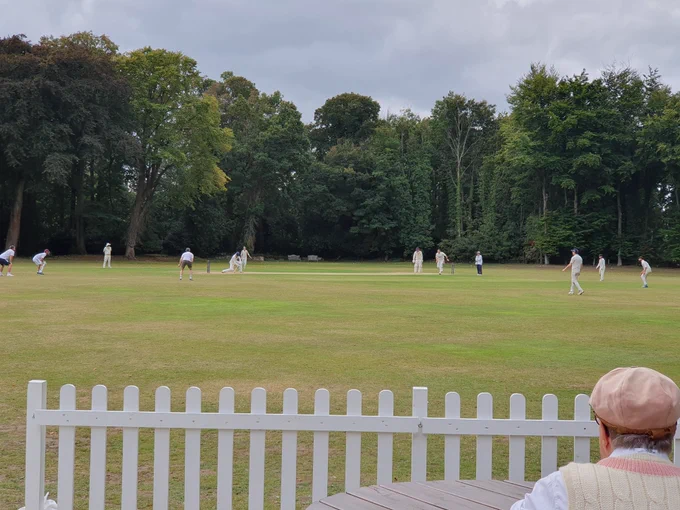 View from the boundary shows cricketers shows slip, gully and other fielders watching on as batter tries to hit ball and as umpire looks on . A white fence and spectating cricketer is seen in the foreground