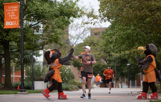 Runner high-fiving a mascot at a Bowling Green State University event, with other mascots nearby, set on a tree-lined campus path.