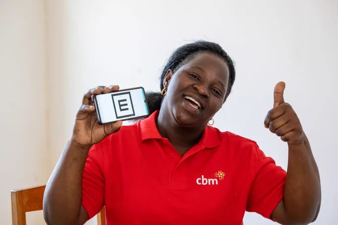 A CBM staff member in a red branded polo demonstrates the Peek Vision software on a smartphone, smiling and giving a thumbs-up gesture.