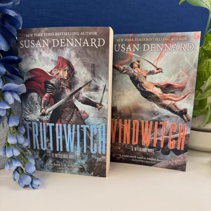 A paperback copy of Truthwitch standing next to a paperback copy of Windwitch, both by Susan Dennard. There is a blue flower next to Truthwitch and a green plant next to Windwitch.