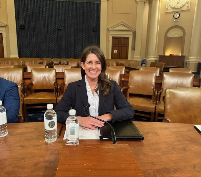 A photo of Denise Hoffman in the committee room, at a table and smiling for the picture.