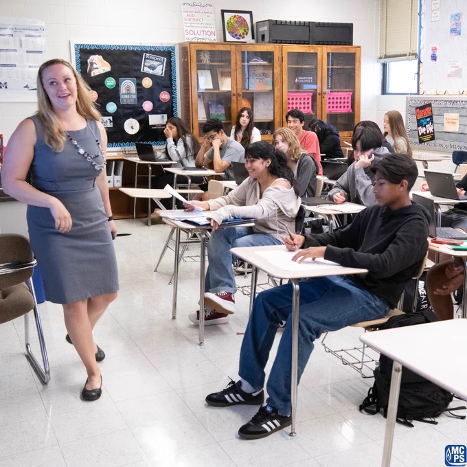 High school students attentively listening to a teacher in a classroom filled with desks and educational posters.